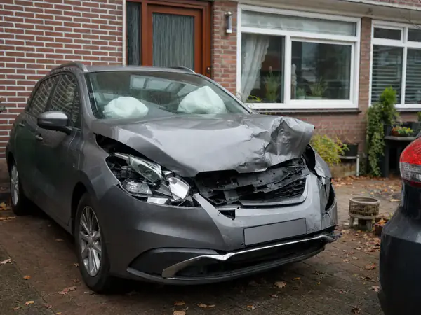 Grijze hatchback met zware frontschade en uitgeklapte airbags op de oprit van een rijtjeshuis