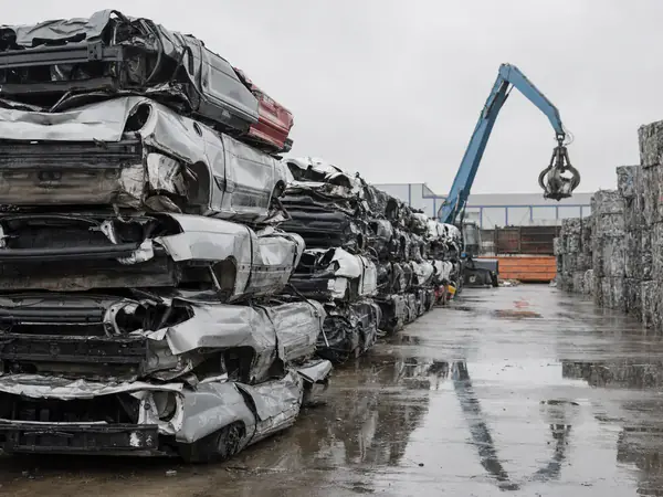 Gestapelde platgedrukte autowrakken op een metaalrecyclingterrein met een kraan op de achtergrond