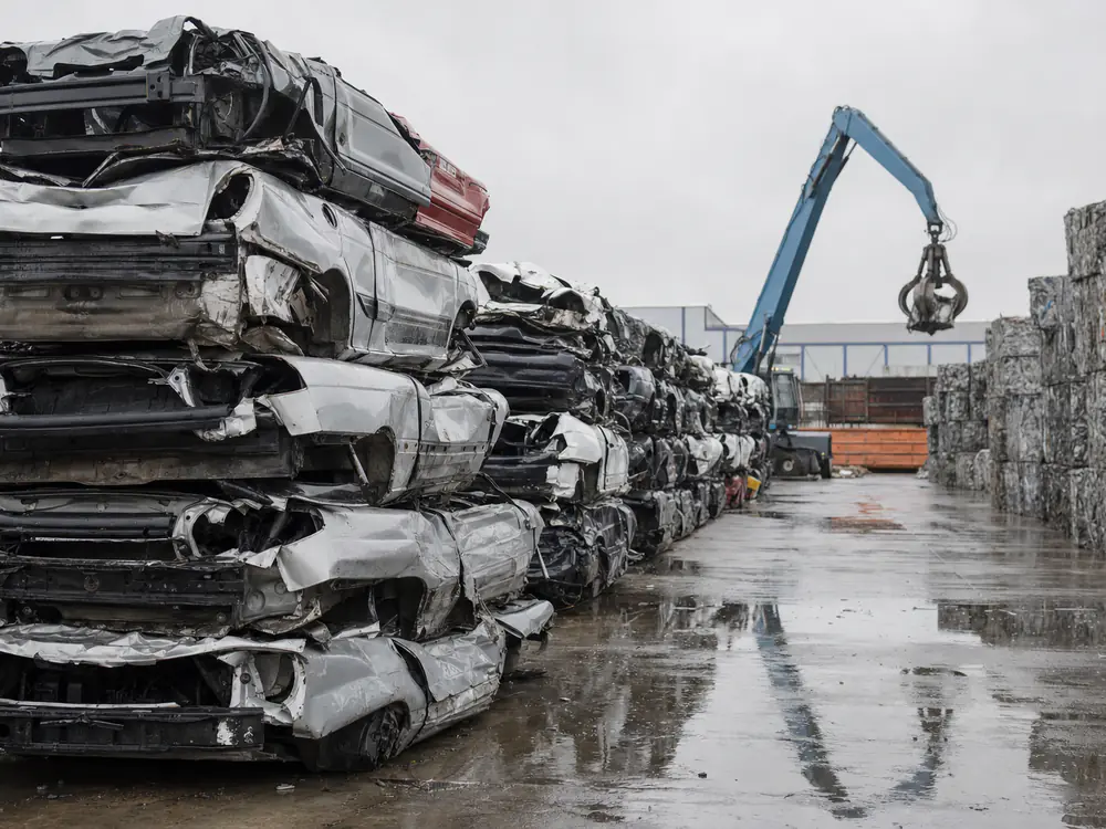 Gestapelde platgedrukte autowrakken op een metaalrecyclingterrein met een kraan op de achtergrond